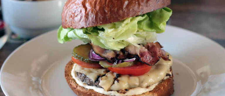 Hamburguesa con pan redondo tostado, lechuga, beicon, queso, pepinillo, tomate y carne de ternera.